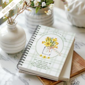 May include: A notebook with a spiral binding and a green plaid design. The cover displays a floral illustration, the text "Best Mummy in the World", and a quote. A vase with flowers is in the background.