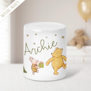 May include: White ceramic money bank with a cartoon illustration of Winnie the Pooh and Piglet. The bank is personalised with the name "Archie" in green script, and has gold stars. Piglet holds a gift. The words "Matching Products" are in the upper left corner.