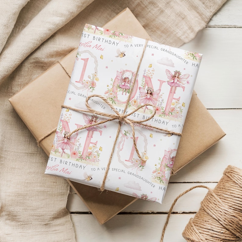 May include: A rectangular gift wrapped in white paper featuring a pink fairy-themed design with the word ONE, floral accents, and text reading 1st Birthday to a very special granddaughter Amelia Mai, tied with a brown twine bow in the center.