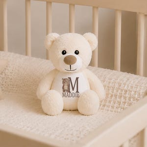 May include: A cream-colored plush teddy bear with a soft, fuzzy texture. The bear features a tan snout, black eyes, and a white patch on its chest. The patch displays a small brown bear illustration, the letter M, and the name MADDIE in dark text.