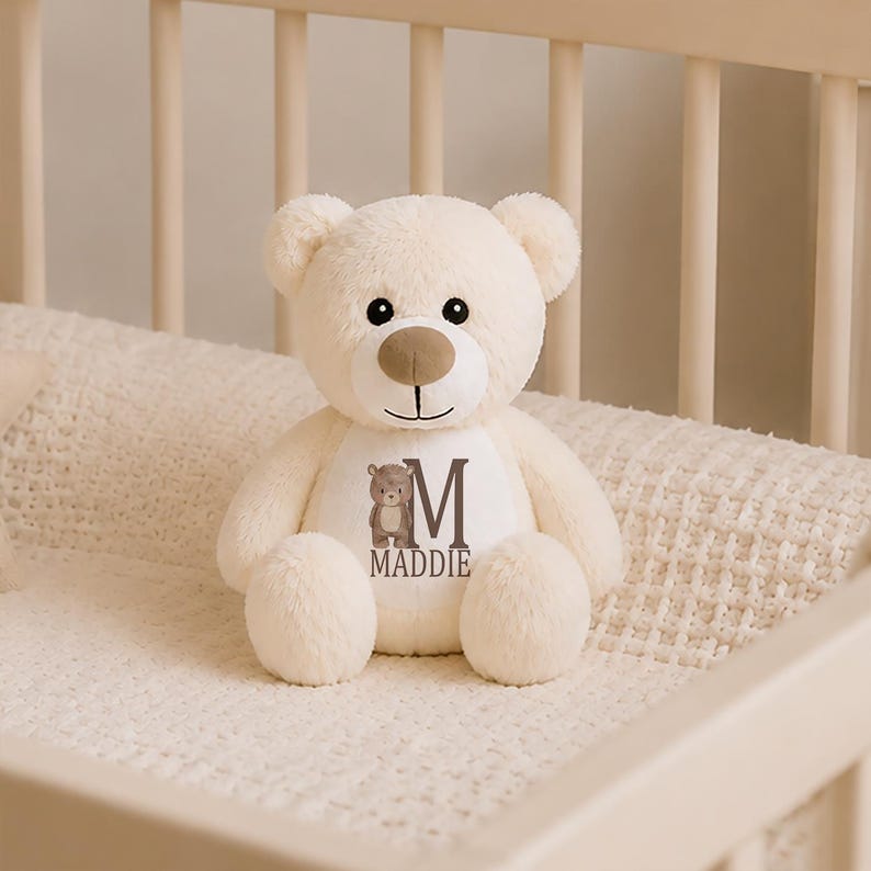 May include: A cream-colored plush teddy bear with a soft, fuzzy texture. The bear features a tan snout, black eyes, and a white patch on its chest. The patch displays a small brown bear illustration, the letter M, and the name MADDIE in dark text.