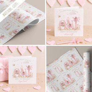 May include: A collection of birthday stationery featuring white greeting cards and wrapping paper with pink fairy illustrations. The designs include the text "ONE" and "HAPPY 1ST BIRTHDAY" alongside floral accents, bees, and small winged fairies.