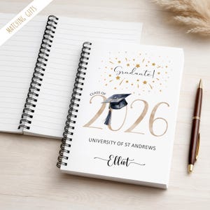 May include: A white spiral-bound notebook with lined pages, featuring a graduation design. The cover includes the text "Graduate!" and "Class of 2026", a graduation cap illustration, and the name "Elliot". A brown pen rests beside the notebook.