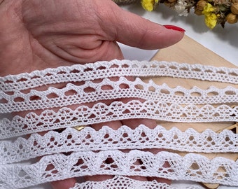May include: A collection of five distinct white lace trim strips featuring various intricate woven patterns, including scalloped edges, geometric cutouts, and floral-inspired motifs, displayed in a layered arrangement for textile projects.