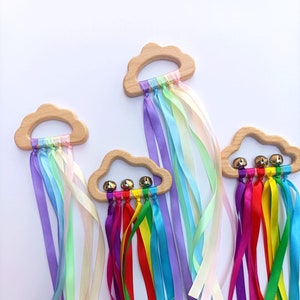 May include: Three wooden cloud-shaped teethers with rainbow-coloured ribbons and bells. The teethers are made of natural wood and have a smooth, rounded shape. The ribbons are tied to the teethers and hang down, creating a playful and colourful sensory experience for babies.