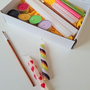 Könnte beinhalten: Eine weiße Box mit Kerzenherstellungszubehör. Die Box enthält weiße Stabkerzen, bunte Teelichter und kleinere Stumpenkerzen in Rosa und Grün. Zwei fertige Kerzen werden gezeigt, eine mit roten Herzen und eine weitere mit einem Regenbogen-Twist-Design. Ein Pinsel ist ebenfalls enthalten.