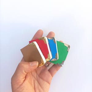 Puede incluir: Cuatro pequeños cuadernos decorativos con detalles dorados y plateados. Los cuadernos son marrones, rojos, azules y verdes. Los cuadernos se sostienen en una mano.