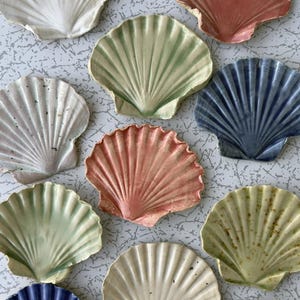 Può includere: Una collezione di piatti in ceramica a forma di conchiglia in vari colori, tra cui bianco, rosa, verde e blu. I piatti presentano un design strutturato e a coste, che ricorda le vere conchiglie. Ideali per servire piccoli snack o come elementi decorativi.
