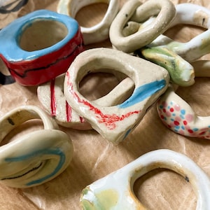 Könnte beinhalten: Eine Sammlung handgefertigter Keramikringe in verschiedenen Formen und Größen. Jeder Ring ist mit leuchtenden Farben bemalt, darunter Rot, Blau und Grün, und liegt auf einer braunen Papieroberfläche. Die Ringe haben ein einzigartiges, künstlerisches Design.