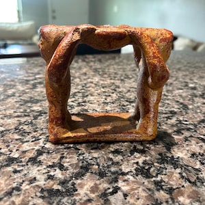 May include: A square, ceramic sculpture in shades of brown and orange. The sculpture features four figures supporting a flat surface, creating an open, rectangular shape. The figures appear to be in a kneeling or crouching position, with their arms raised.