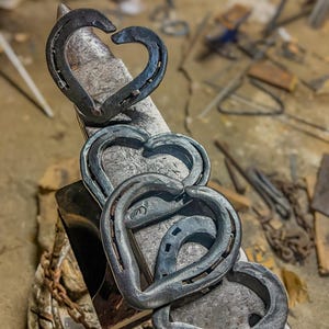 K&ouml;nnte beinhalten: Handgeschmiedete Metallherzen aus Hufeisen. Die Herzen sind in einer Kette angeordnet, wobei jedes Hufeisen eine Herzform bildet. Das Metall hat eine dunkle, strukturierte Oberfl&auml;che, und die Herzen werden auf einem Amboss in einer Werkstatt gezeigt.