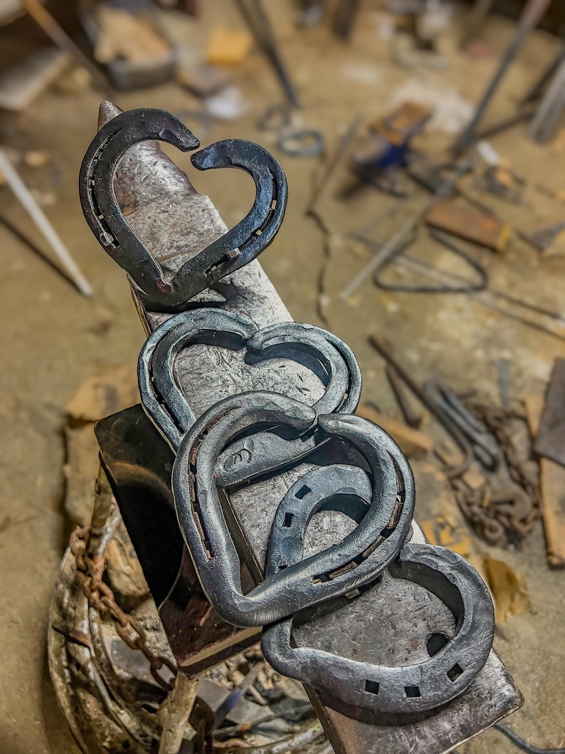 K&ouml;nnte beinhalten: Handgeschmiedete Metallherzen aus Hufeisen. Die Herzen sind in einer Kette angeordnet, wobei jedes Hufeisen eine Herzform bildet. Das Metall hat eine dunkle, strukturierte Oberfl&auml;che, und die Herzen werden auf einem Amboss in einer Werkstatt gezeigt.