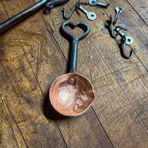 Op de afbeelding: Een handgesmede koperen schep met een donker, getextureerd ijzeren handvat in de vorm van een hart aan de bovenkant. De schep heeft een gehamerde afwerking en een enkele klinknagel. Diverse andere ijzeren haken en gereedschappen liggen eromheen.