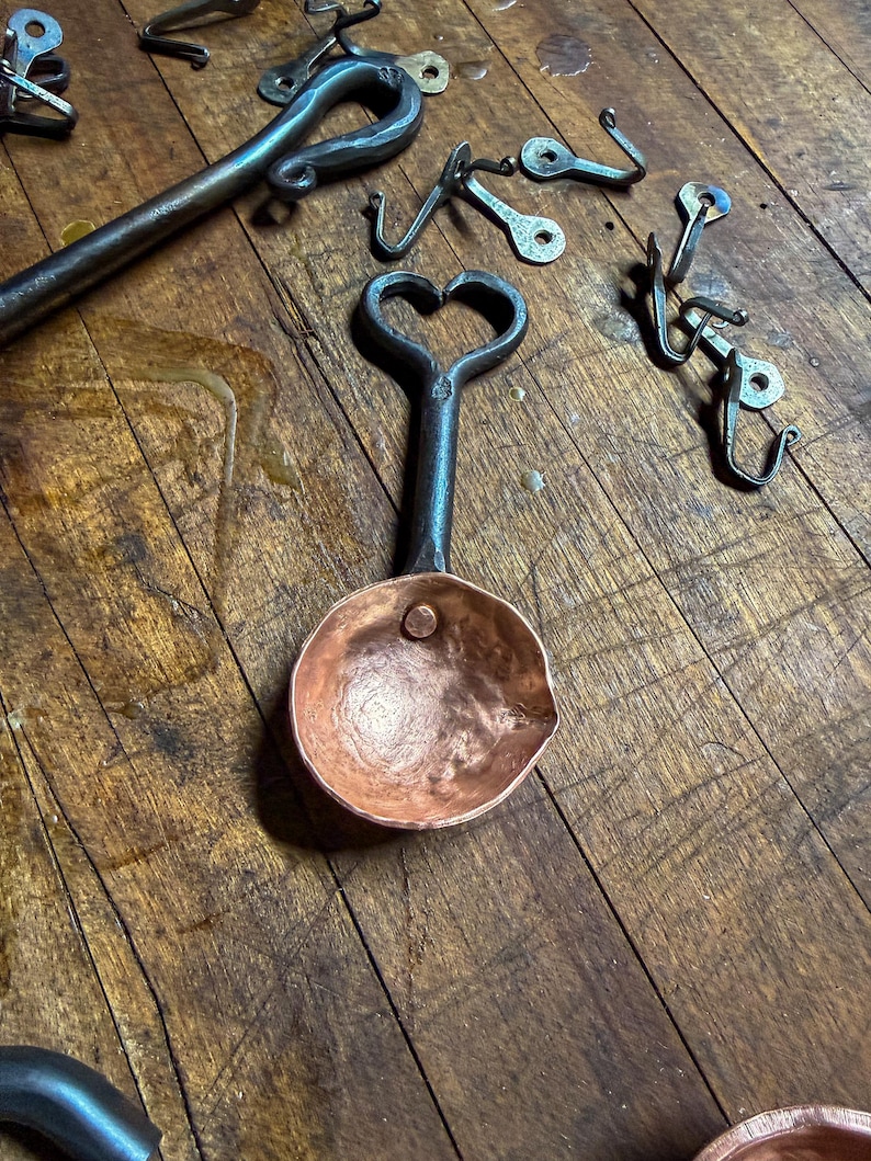 Op de afbeelding: Een handgesmede koperen schep met een donker, getextureerd ijzeren handvat in de vorm van een hart aan de bovenkant. De schep heeft een gehamerde afwerking en een enkele klinknagel. Diverse andere ijzeren haken en gereedschappen liggen eromheen.