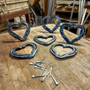 K&ouml;nnte beinhalten: Sechs handgefertigte herzf&ouml;rmige Metallobjekte aus Hufeisen, angeordnet auf einer Holzoberfl&auml;che. Die Herzen variieren in der Gr&ouml;&szlig;e und haben eine dunkle, metallische Farbe. Mehrere Metalln&auml;gel sind in der N&auml;he der Herzen verstreut.