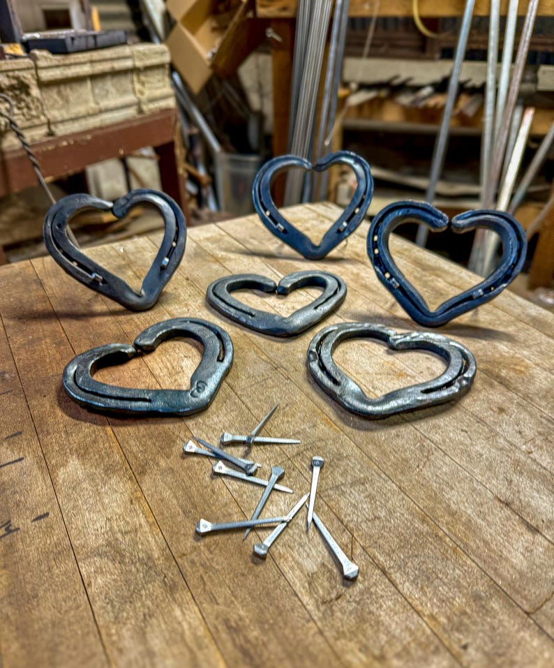 K&ouml;nnte beinhalten: Sechs handgefertigte herzf&ouml;rmige Metallobjekte aus Hufeisen, angeordnet auf einer Holzoberfl&auml;che. Die Herzen variieren in der Gr&ouml;&szlig;e und haben eine dunkle, metallische Farbe. Mehrere Metalln&auml;gel sind in der N&auml;he der Herzen verstreut.