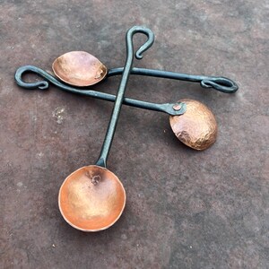 Op de afbeelding: Twee handgesmede lepels met ronde, gehamerde koperen kommen bevestigd aan lange, donkergrijze ijzeren stelen. Elke steel eindigt in een decoratieve gekrulde lus. De lepels zijn in een gekruiste opstelling op een plat oppervlak geplaatst.