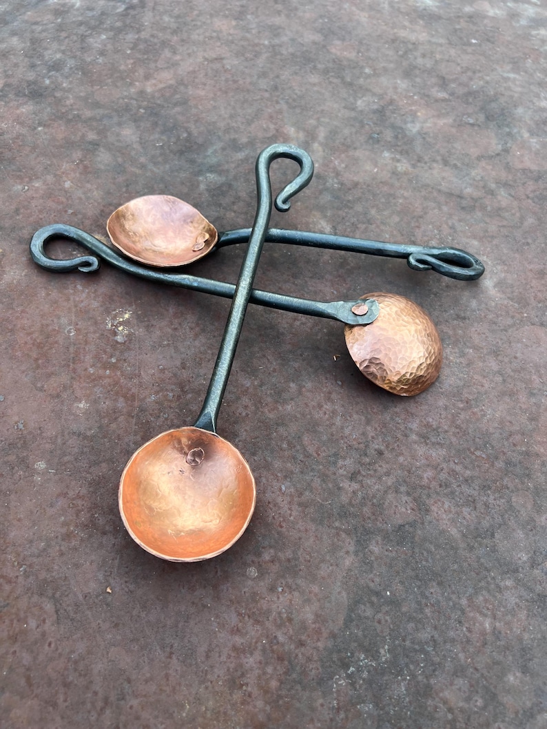 Op de afbeelding: Twee handgesmede lepels met ronde, gehamerde koperen kommen bevestigd aan lange, donkergrijze ijzeren stelen. Elke steel eindigt in een decoratieve gekrulde lus. De lepels zijn in een gekruiste opstelling op een plat oppervlak geplaatst.
