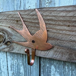 Op de afbeelding: Een koperkleurige metalen wandhaak in de vorm van een zwaluw met gegraveerde vleugeldetails. De vogel is gemonteerd op een verweerde, horizontale houten plank met een enkele donkere schroef in het midden van het lichaam en een gebogen haak.
