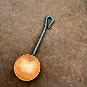 Op de afbeelding: Een handgesmede metalen pollepel met een ronde, gehamerde koperen kom bevestigd aan een lange, donkere ijzeren steel die eindigt in een decoratieve gekrulde haak. De kom heeft een gladde, metalen afwerking met een enkele kleine klinknagel.