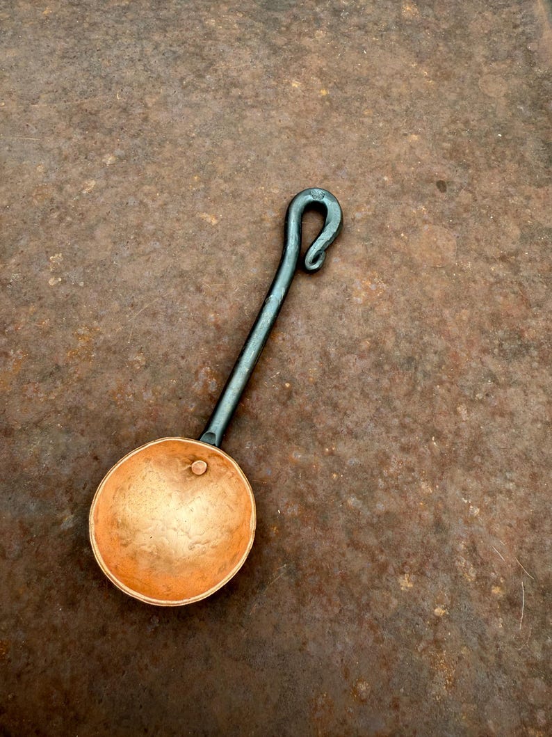 Op de afbeelding: Een handgesmede metalen pollepel met een ronde, gehamerde koperen kom bevestigd aan een lange, donkere ijzeren steel die eindigt in een decoratieve gekrulde haak. De kom heeft een gladde, metalen afwerking met een enkele kleine klinknagel.
