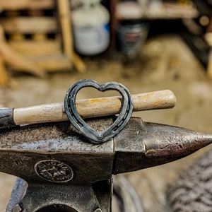 K&ouml;nnte beinhalten: Ein herzf&ouml;rmiges Hufeisen liegt auf einem Schmiedeamboss, mit einem Hammer im Hintergrund. Das Hufeisen ist aus dunklem Metall gefertigt und der Amboss ist dunkelgrau. Der Hammer hat einen Holzgriff. Der Hintergrund ist unscharf.
