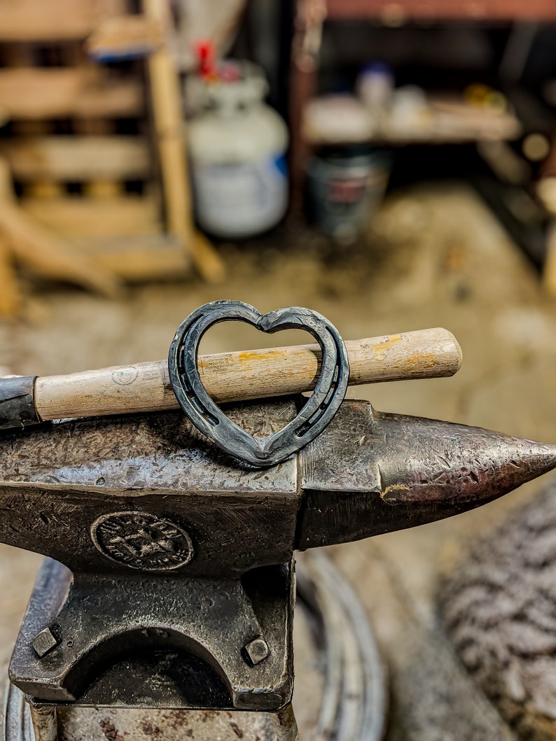 K&ouml;nnte beinhalten: Ein herzf&ouml;rmiges Hufeisen liegt auf einem Schmiedeamboss, mit einem Hammer im Hintergrund. Das Hufeisen ist aus dunklem Metall gefertigt und der Amboss ist dunkelgrau. Der Hammer hat einen Holzgriff. Der Hintergrund ist unscharf.