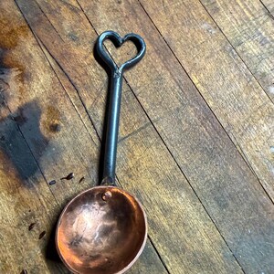 Op de afbeelding: Een kleine metalen maatlepel met een ronde, gepolijste koperen kom bevestigd aan een donker, gesmeed ijzeren handvat. Het handvat eindigt in een decoratieve open hartvormige lus aan de bovenkant, wat een contrast vormt tussen koper en ijzer.