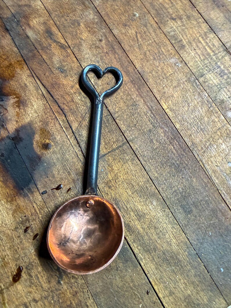 Op de afbeelding: Een kleine metalen maatlepel met een ronde, gepolijste koperen kom bevestigd aan een donker, gesmeed ijzeren handvat. Het handvat eindigt in een decoratieve open hartvormige lus aan de bovenkant, wat een contrast vormt tussen koper en ijzer.