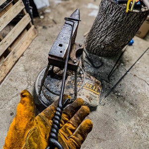 Puede incluir: Una herramienta de herrero de metal negro forjada a mano, con un mango retorcido y un lazo circular. La herramienta se sostiene con un guante de trabajo de cuero amarillo. Un yunque y una bolsa de hormigón están en el fondo.