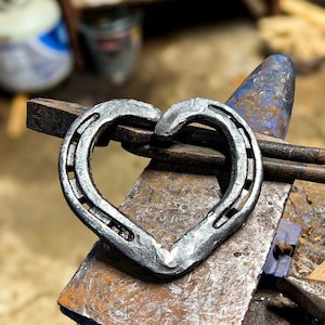 K&ouml;nnte beinhalten: Ein handgeschmiedetes Herz aus Hufeisen, das auf einem Amboss liegt. Die Herzform entsteht durch das Zusammenf&uuml;gen der gebogenen Enden der Hufeisen. Das Metall hat eine dunkle, strukturierte Oberfl&auml;che, der Amboss ist rostig.