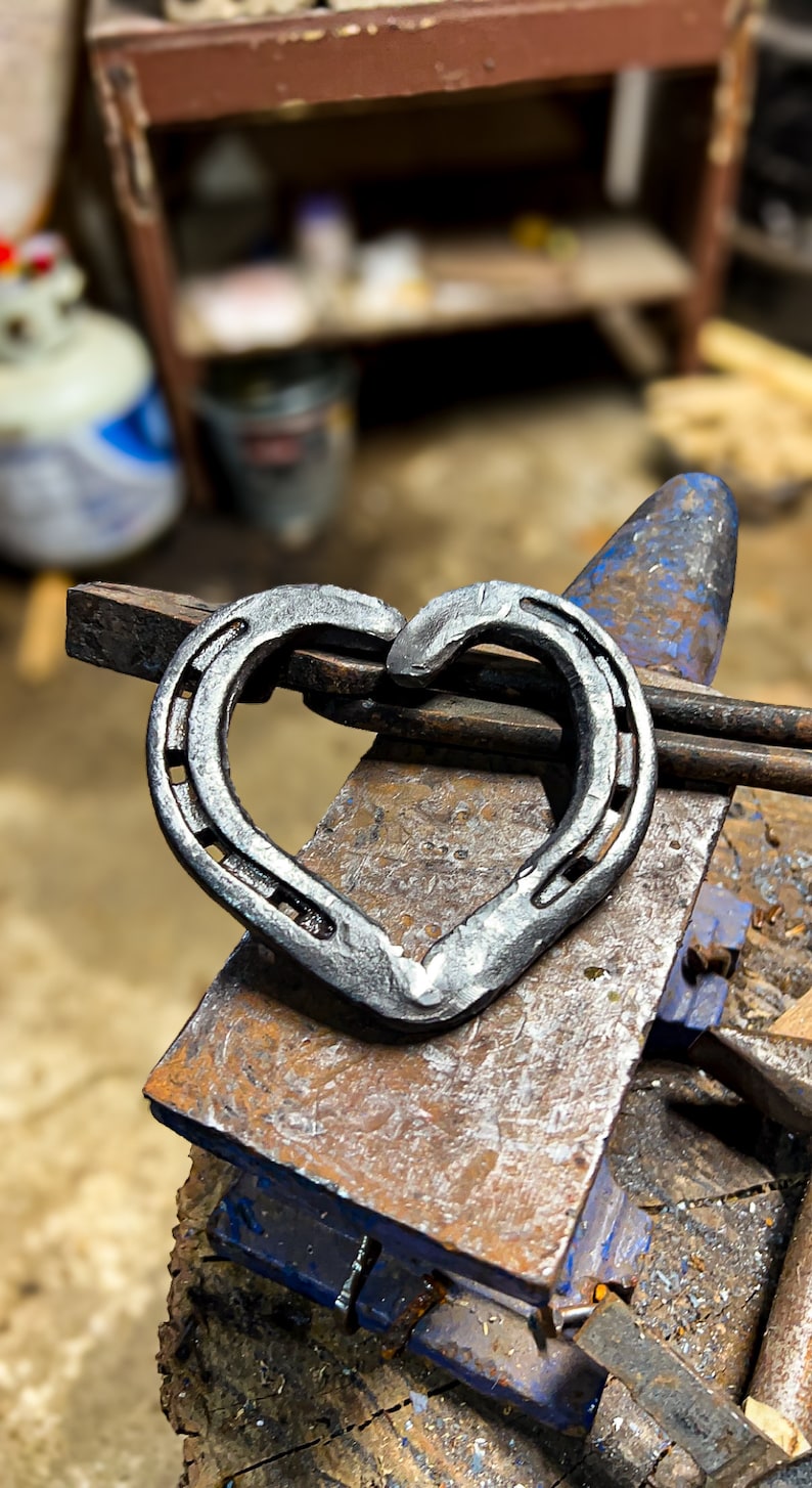 K&ouml;nnte beinhalten: Ein handgeschmiedetes Herz aus Hufeisen, das auf einem Amboss liegt. Die Herzform entsteht durch das Zusammenf&uuml;gen der gebogenen Enden der Hufeisen. Das Metall hat eine dunkle, strukturierte Oberfl&auml;che, der Amboss ist rostig.