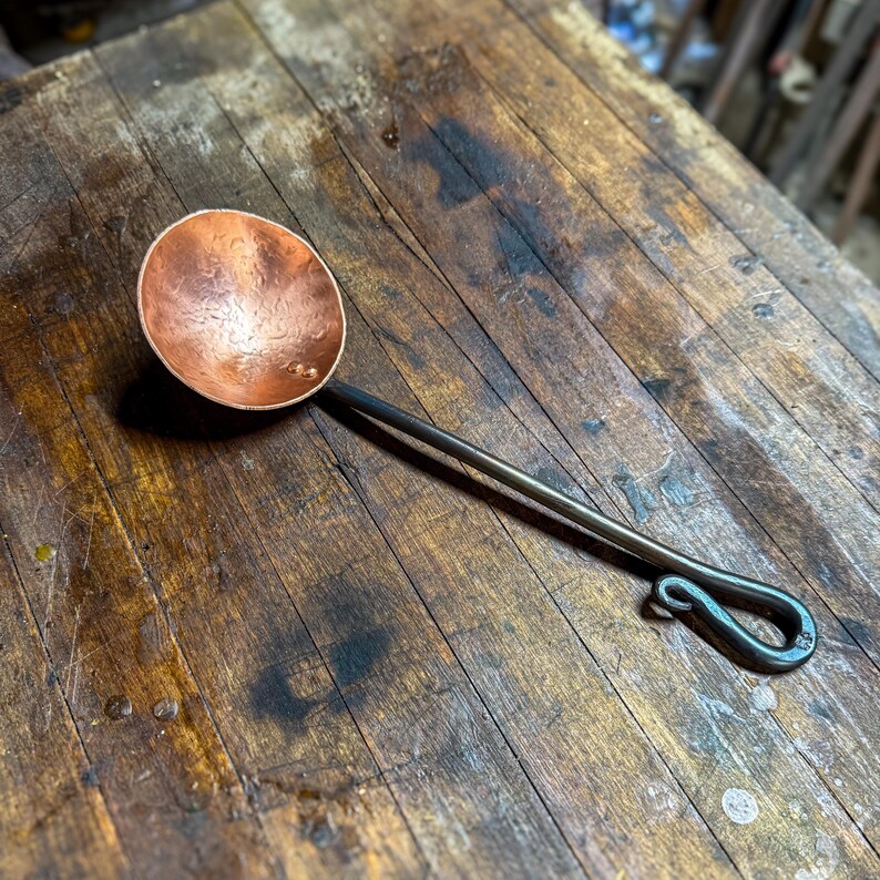 Op de afbeelding: Een handgesmede pollepel met een ronde, gehamerde koperen kom bevestigd aan een lange, donkere ijzeren steel. De steel eindigt in een decoratieve, gekrulde lus, wat zorgt voor een functionele grip voor dit keukengerei.