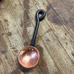 Op de afbeelding: Een kleine, ronde, gehamerde koperen schep met een donker, gesmeed ijzeren handvat. Het handvat heeft een lusvormig uiteinde om op te hangen. Twee kleine koperen klinknagels bevestigen de kom van de schep aan het handvat, een functioneel keukengerei.