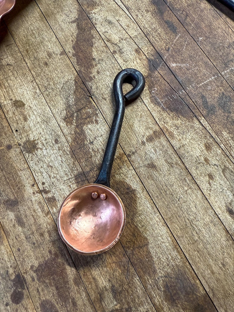 Op de afbeelding: Een kleine, ronde, gehamerde koperen schep met een donker, gesmeed ijzeren handvat. Het handvat heeft een lusvormig uiteinde om op te hangen. Twee kleine koperen klinknagels bevestigen de kom van de schep aan het handvat, een functioneel keukengerei.