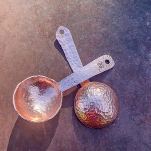 May include: Two hammered copper measuring spoons, one with a handle and one without. The spoons are on a textured gray surface.