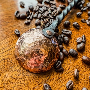 Op de afbeelding: Een koperkleurige metalen koffieschep met een ronde, gehamerde kom en een gedraaide zwarte metalen steel. De schep is met een enkele klinknagel aan de steel bevestigd en wordt omringd door verspreide donkere koffiebonen op een houten ondergrond.