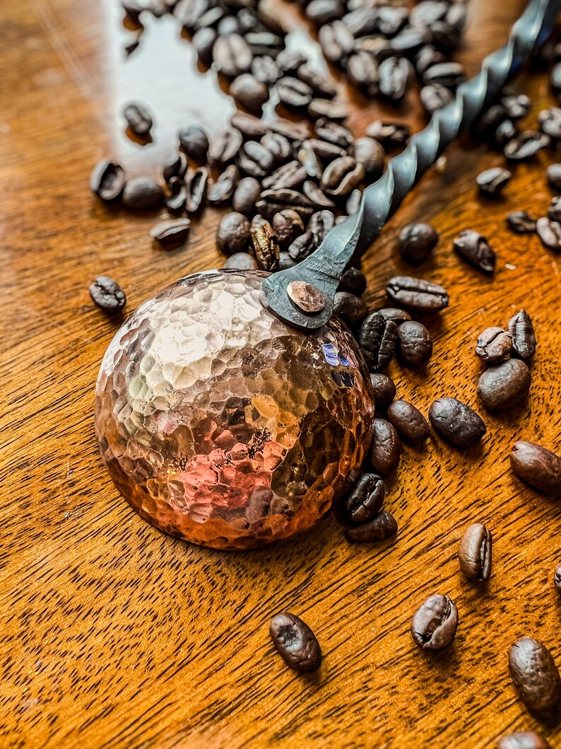Op de afbeelding: Een koperkleurige metalen koffieschep met een ronde, gehamerde kom en een gedraaide zwarte metalen steel. De schep is met een enkele klinknagel aan de steel bevestigd en wordt omringd door verspreide donkere koffiebonen op een houten ondergrond.