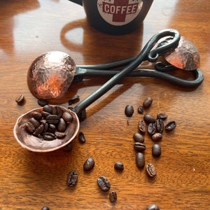 Op de afbeelding: Twee handgesmede metalen koffiescheppen met gehamerde koperen kommen en donkere, gedraaide ijzeren handvatten. E&eacute;n schep is gevuld met geroosterde koffiebonen, terwijl de andere ernaast ligt. Een zwarte mok met het woord COFFEE is op de achtergrond zichtbaar.