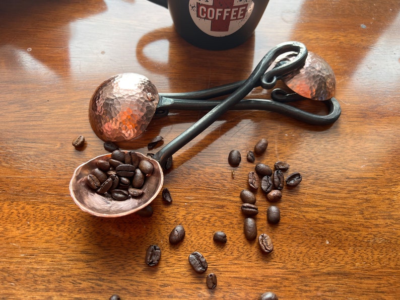 Op de afbeelding: Twee handgesmede metalen koffiescheppen met gehamerde koperen kommen en donkere, gedraaide ijzeren handvatten. E&eacute;n schep is gevuld met geroosterde koffiebonen, terwijl de andere ernaast ligt. Een zwarte mok met het woord COFFEE is op de achtergrond zichtbaar.