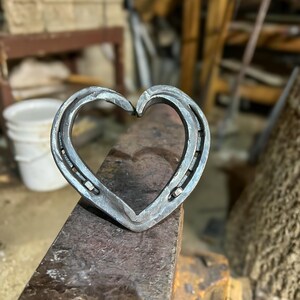K&ouml;nnte beinhalten: Ein handgefertigtes Herz aus Hufeisen, platziert auf einem dunklen, strukturierten Amboss. Das Metallherz zeigt einen rustikalen, industriellen Stil. Geeignet als Dekoration oder einzigartiges Geschenk.