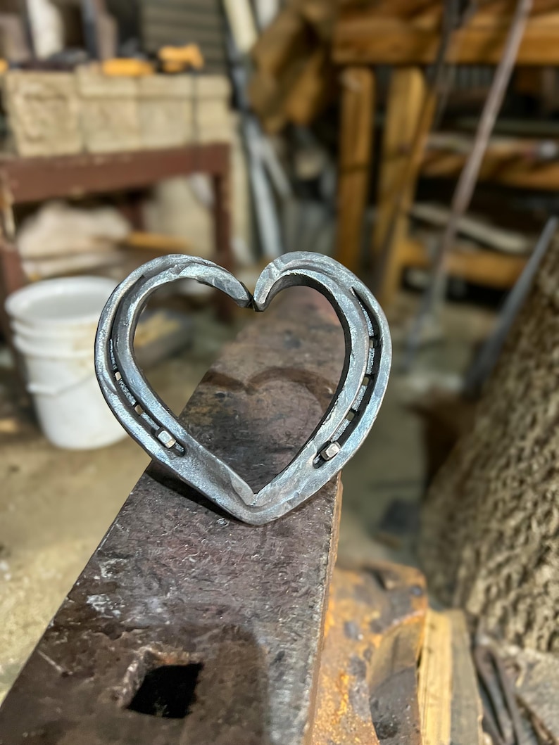 K&ouml;nnte beinhalten: Ein handgefertigtes Herz aus Hufeisen, platziert auf einem dunklen, strukturierten Amboss. Das Metallherz zeigt einen rustikalen, industriellen Stil. Geeignet als Dekoration oder einzigartiges Geschenk.