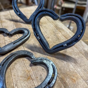 K&ouml;nnte beinhalten: Mehrere handgefertigte Metallherzskulpturen aus Hufeisen. Die Herzen sind dunkelgrau und scheinen aus Eisen oder Stahl zu sein. Sie sind auf einer Holzoberfl&auml;che angeordnet, wobei einige im Fokus und andere im Hintergrund unscharf sind. Das rustikale Design deutet auf ein handgefertigtes Produkt hin.