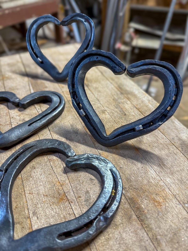 K&ouml;nnte beinhalten: Mehrere handgefertigte Metallherzskulpturen aus Hufeisen. Die Herzen sind dunkelgrau und scheinen aus Eisen oder Stahl zu sein. Sie sind auf einer Holzoberfl&auml;che angeordnet, wobei einige im Fokus und andere im Hintergrund unscharf sind. Das rustikale Design deutet auf ein handgefertigtes Produkt hin.