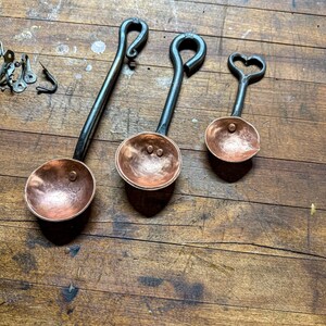 Op de afbeelding: Drie handgesmede maatlepels met gehamerde koperen kommen en donkere ijzeren handvatten. De handvatten hebben opvallende lusontwerpen, waaronder een hartvormig uiteinde aan de kleinste lepel. De koperen oppervlakken hebben een textuur.
