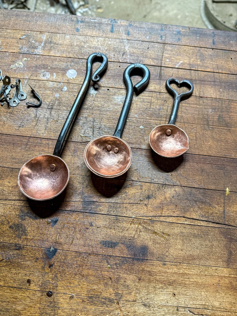 Op de afbeelding: Drie handgesmede maatlepels met gehamerde koperen kommen en donkere ijzeren handvatten. De handvatten hebben opvallende lusontwerpen, waaronder een hartvormig uiteinde aan de kleinste lepel. De koperen oppervlakken hebben een textuur.
