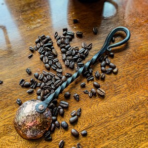Op de afbeelding: Een handgesmede koffieschep met een ronde, gehamerde koperen kom bevestigd aan een gedraaide zwarte metalen steel die eindigt in een decoratieve lus. De schep is omringd door verspreide geroosterde koffiebonen op een houten ondergrond.