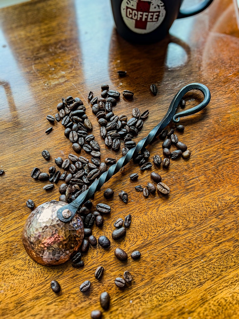 Op de afbeelding: Een handgesmede koffieschep met een ronde, gehamerde koperen kom bevestigd aan een gedraaide zwarte metalen steel die eindigt in een decoratieve lus. De schep is omringd door verspreide geroosterde koffiebonen op een houten ondergrond.