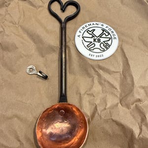 Op de afbeelding: Een handgesmede metalen pollepel met een koperen kom en een lange zwarte steel die eindigt in een hartvormige lus. Daarnaast liggen een kleine metalen haak en een ronde witte sticker met de tekst A FIREMAN'S FORGE KB EST 2022.