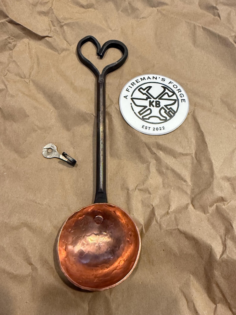 Op de afbeelding: Een handgesmede metalen pollepel met een koperen kom en een lange zwarte steel die eindigt in een hartvormige lus. Daarnaast liggen een kleine metalen haak en een ronde witte sticker met de tekst A FIREMAN'S FORGE KB EST 2022.